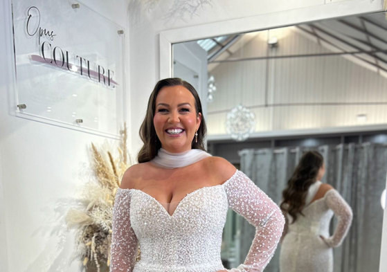 smiling dark-haired model wears off the shoulder long sleeve shimmery wedding dress and a tulle neck scarf