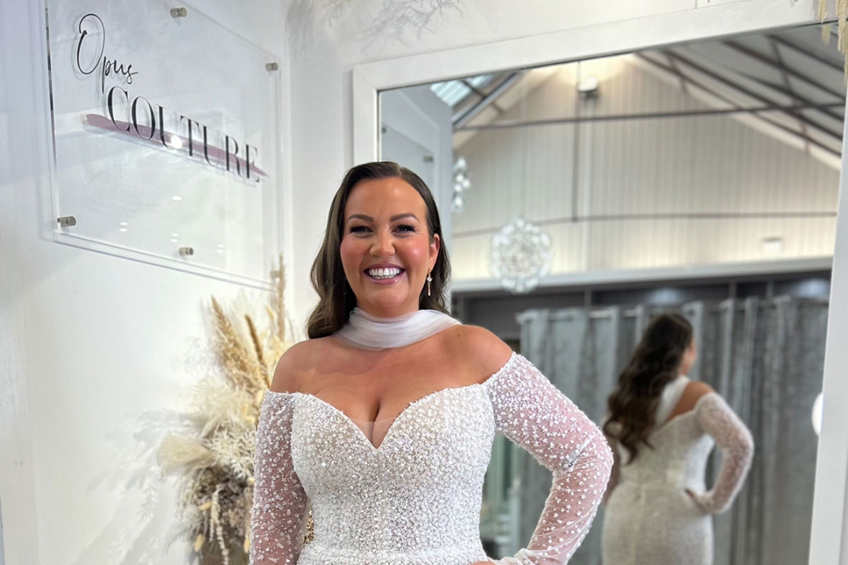 smiling dark-haired model wears off the shoulder long sleeve shimmery wedding dress and a tulle neck scarf