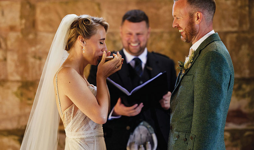 A bride imbibing a drink from the quaich during a wedding ceremony as happy groom and officiant smile
