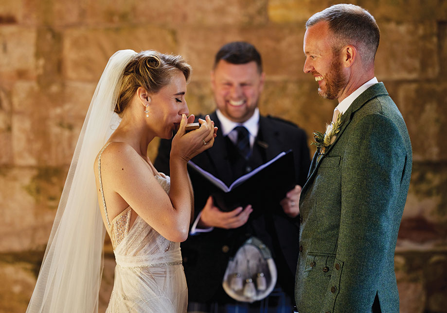 A bride imbibing a drink from the quaich during a wedding ceremony as happy groom and officiant smile