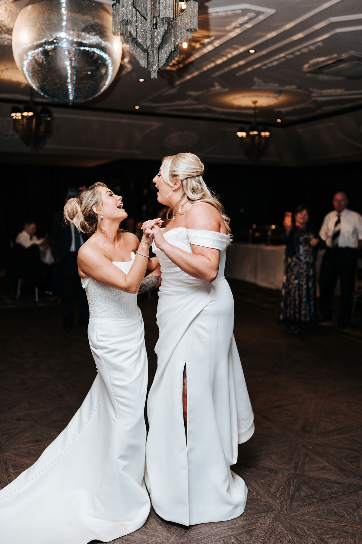 Two brides sharing their first dance beneath a disco ball and chandelier during the evening celebration