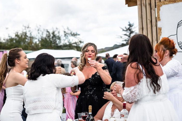 A Bride Wearing A Black Wedding Dress Holds A Small Shot Glass While A Group Of People Wearing White Toast Her