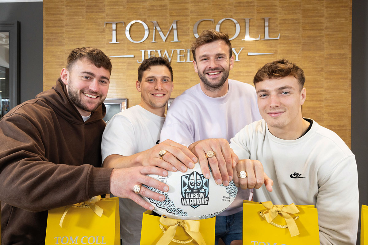 four men stand huddled together in front of Tom Coll Jewellery sign on wall, all reaching out to hold a 'Glasgow Warriors' rugby ball and showing off matching gold rings 