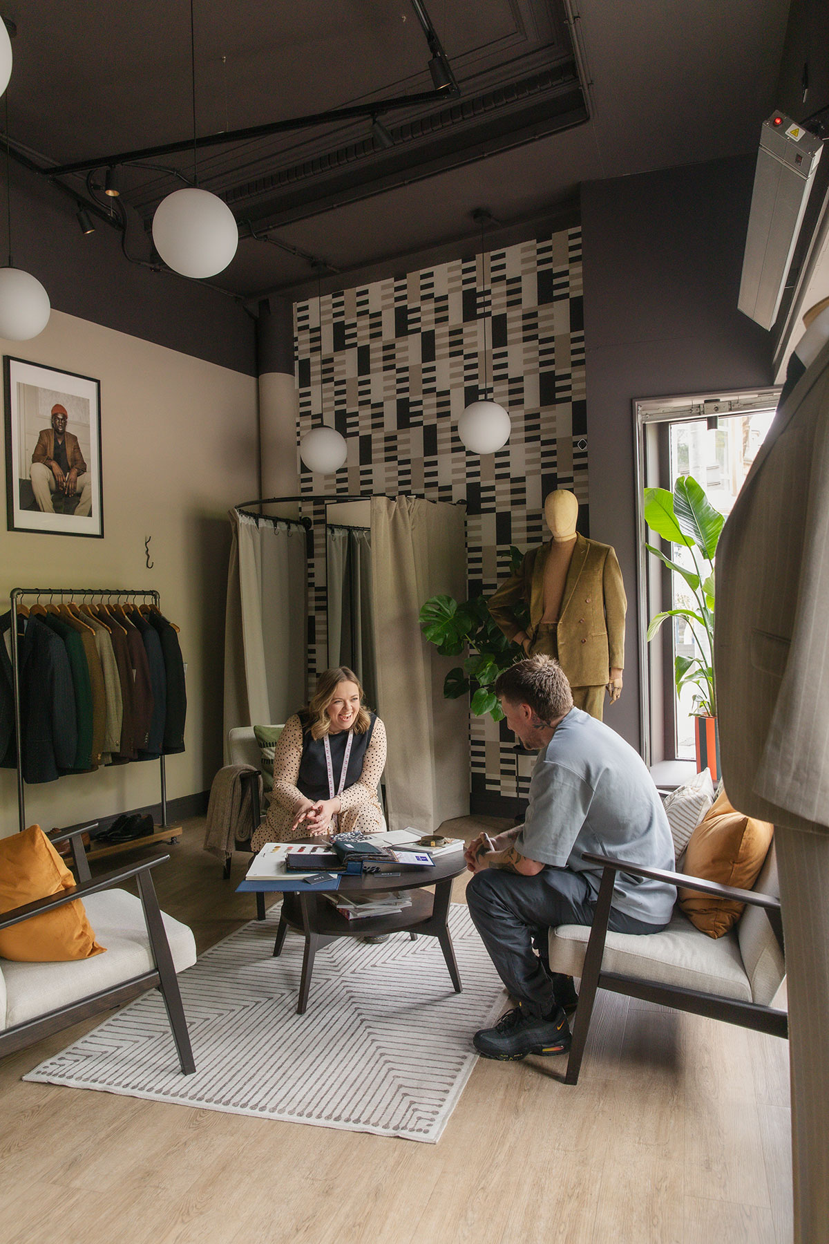 a smiling woman sits across from a man within a clothing store, where they are both sat on cushioned lounge chairs with a stack of papers and books on the coffee table in front of them and jackets and suits displayed on rails and mannequins around them