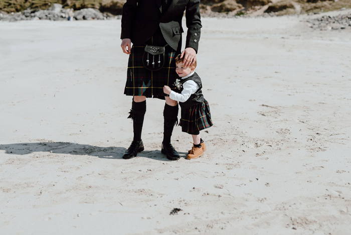 Groom's son holds his leg as they stand on beach wearing kilts