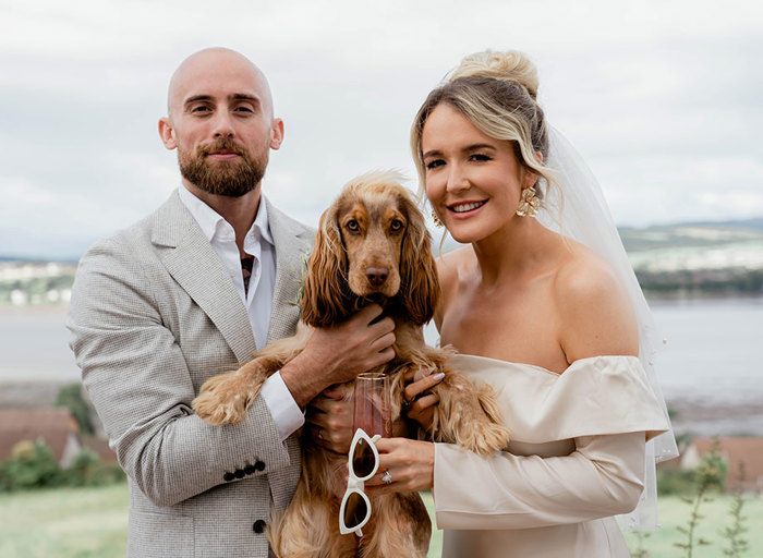 a bride and groom posing with a red spaniel dog with river view in background