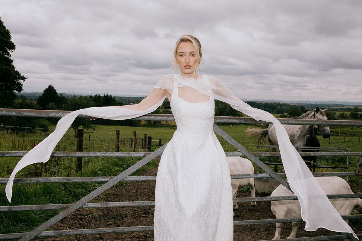 A bride in a white textured gown poses in front of a metal farm gate, with goats and horses in the background and open countryside beyond.