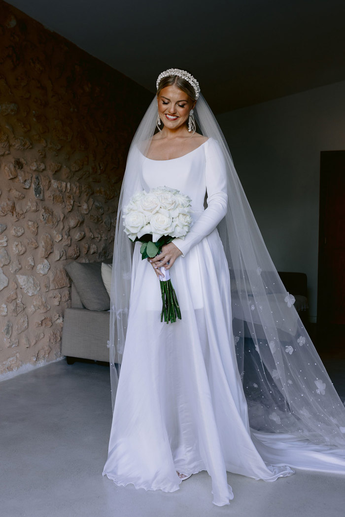 bride in long sleeve mini wedding dress with sheer overskirt and floral applique veil stands and looks down at the bouquet of roses in her hands