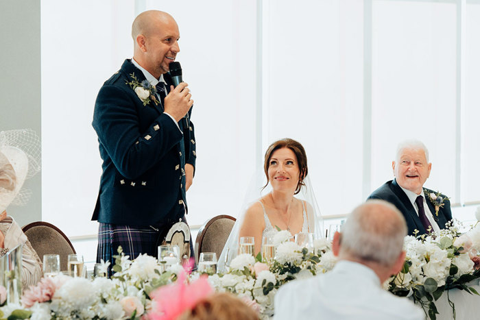 a bride and wedding guests look towards a groom wearing a tartan plaid holding a microphone during his wedding speech