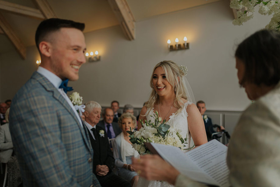 A Smiling Bride And Groom With Celebrant Reading Notes During A Wedding Ceremony As Guests Look On