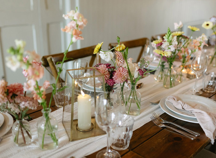 bud vases and candles line the middle of white table runner on a wooden dining table