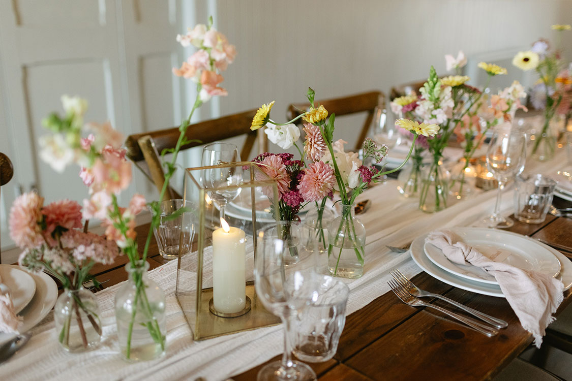 bud vases and candles line the middle of white table runner on a wooden dining table