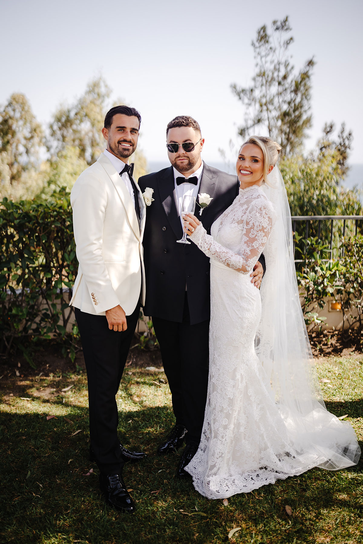 Bride and groom pose with guest in black tuxedo at outdoor reception