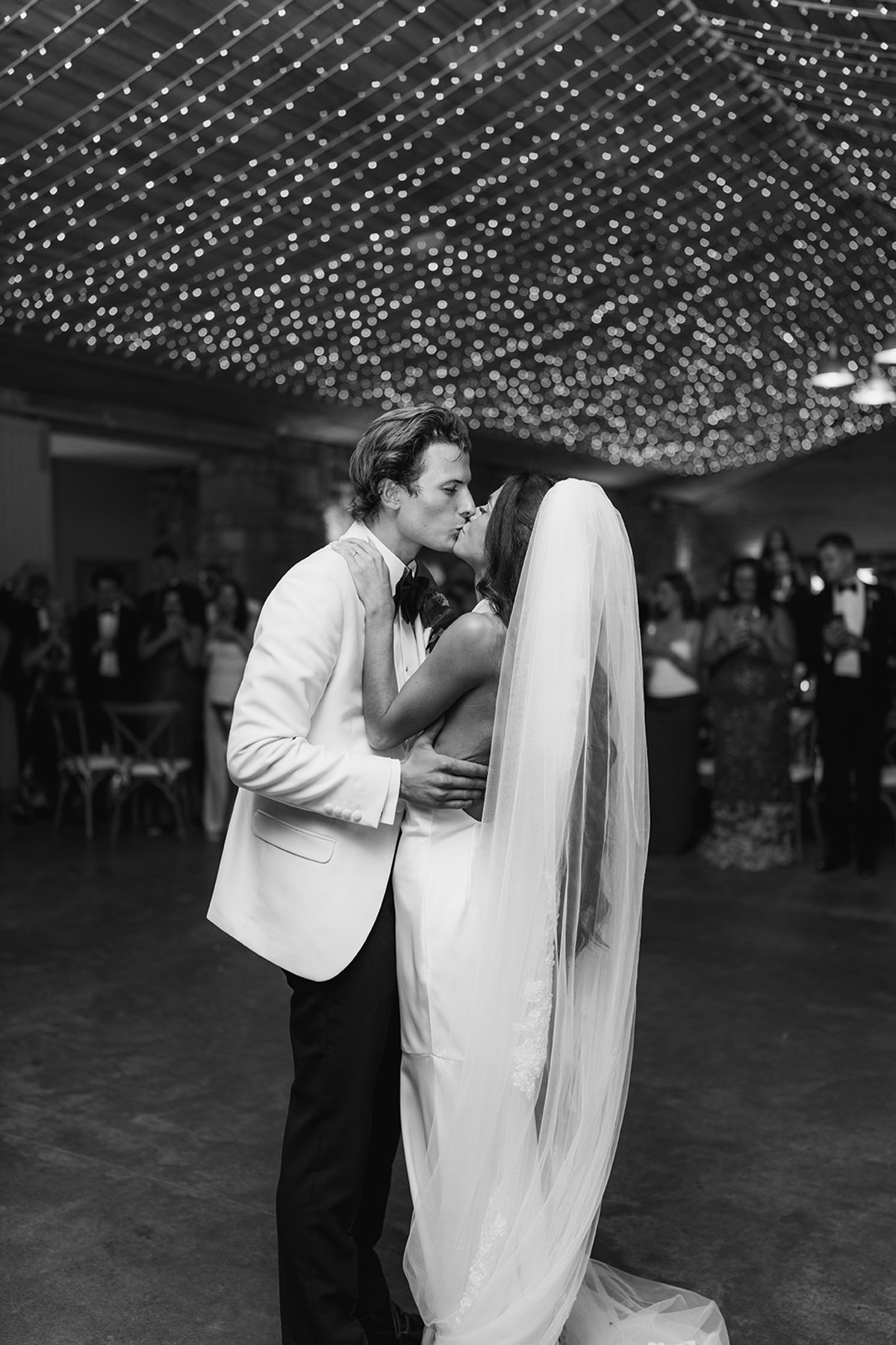 Black and white first dance kiss under fairy lights in rustic wedding reception space
