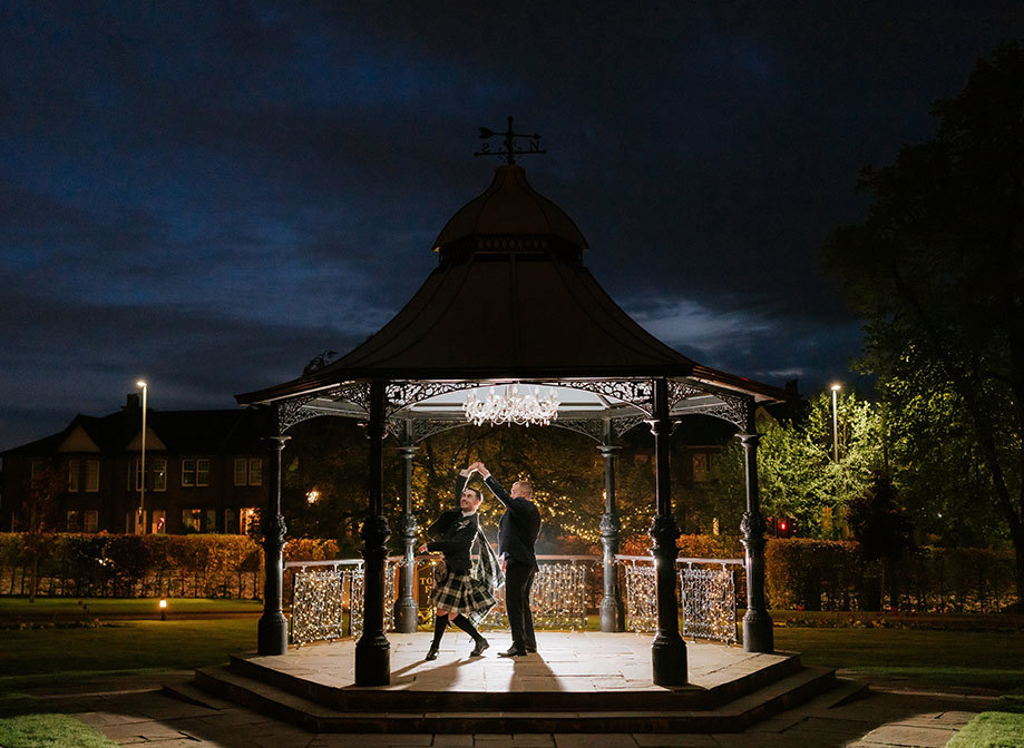 two grooms dancing in an old Victorian bandstand at night time with chandelier illuminating them