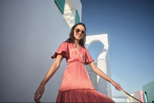 girl smiling wearing a coral dress with floaty sleeves in front of a white wall in the sunshine