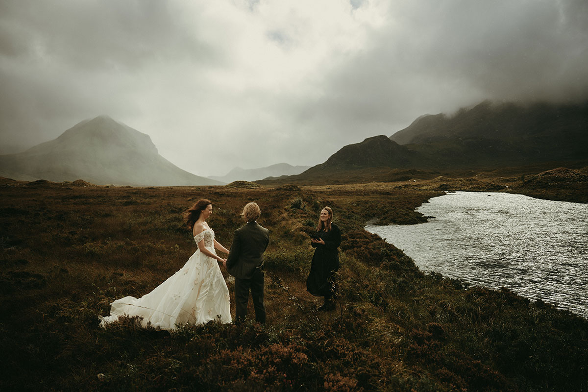 Couple stand hand in hand beside a loch during a remote outdoor wedding ceremony surrounded by dramatic Highland scenery