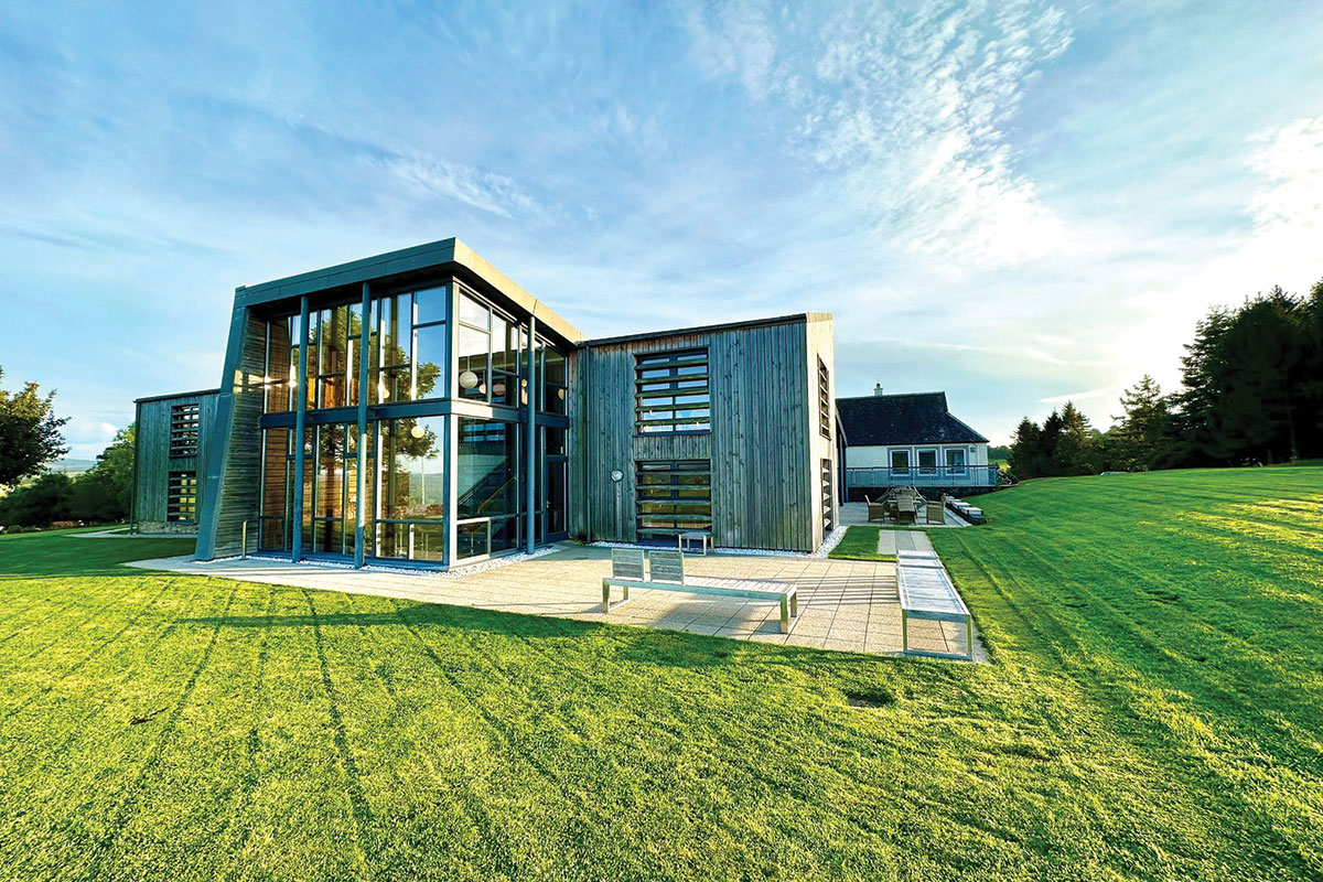 outside view of a large building with floor to ceiling windows and wooden walls, surrounded by very green grass on a blue sky day