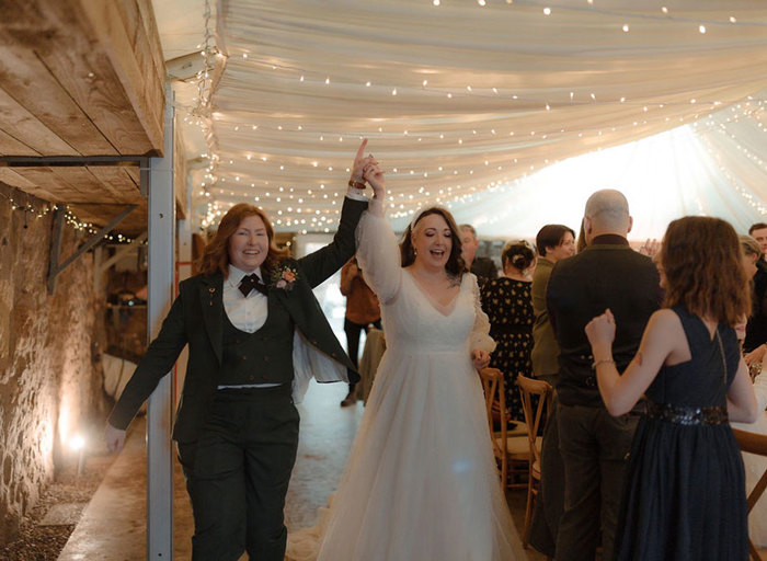 two brides cheer and raise their hands in the air as they enter a room full of wedding guests. There is a draped canvas ceiling with rows of fairy lights above them and a stone wall on the left