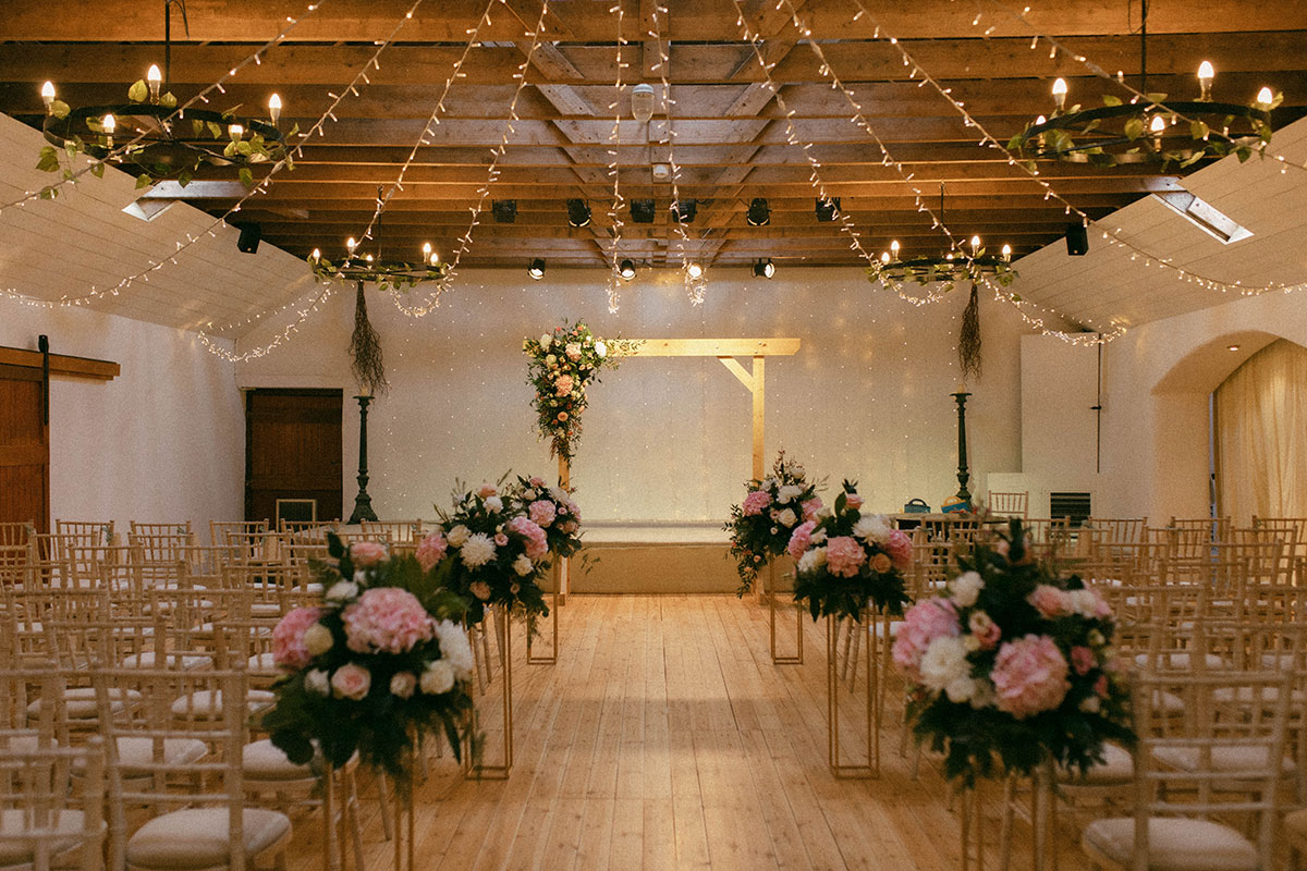 Aswanley's ballroom set up for a wedding ceremony with rows of wooden chairs, pink and white floral arrangements, a wooden ceremony arch and fairy lights suspended from exposed ceiling beams