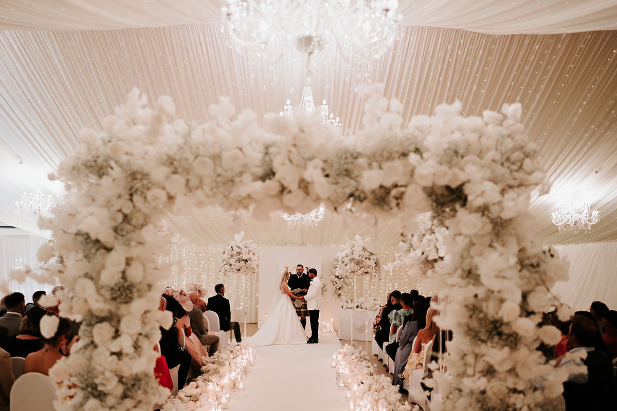 Wedding ceremony at Ingliston Country Club framed by floral arch with candlelit aisle and seated guests