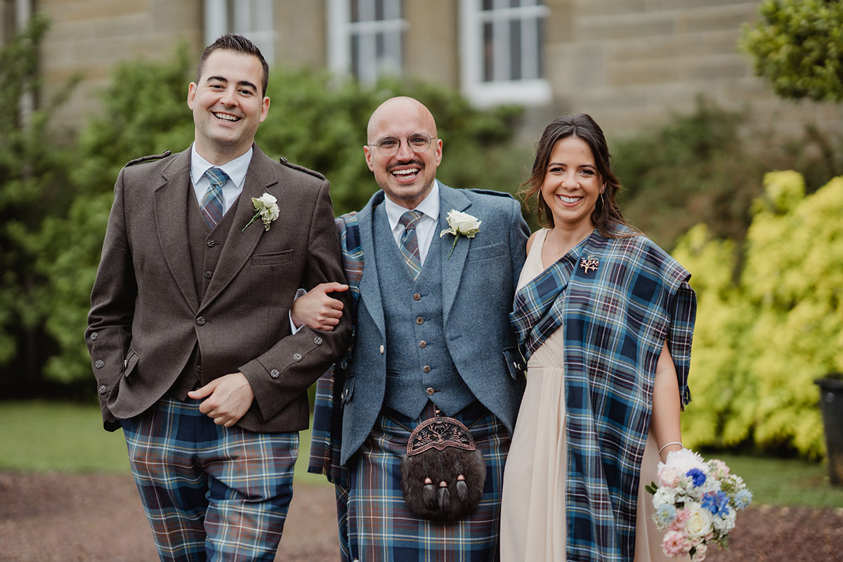 A groom stands arm in arm with two wedding party members outside the venue, all wearing matching tartan elements in shades of blue and brown