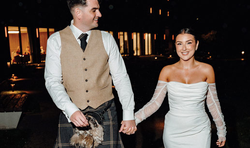 bride in strapless dress with detached tulle sleeves holds hand of tall groom in beige and dark green kilt outfit