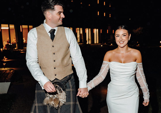 bride in strapless dress with detached tulle sleeves holds hand of tall groom in beige and dark green kilt outfit