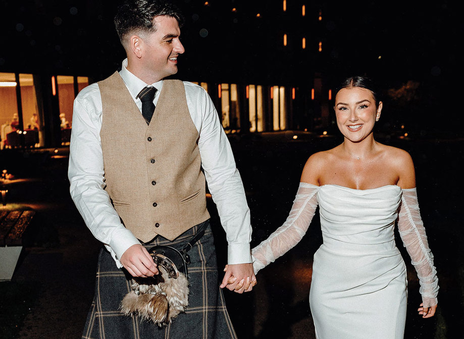 bride in strapless dress with detached tulle sleeves holds hand of tall groom in beige and dark green kilt outfit