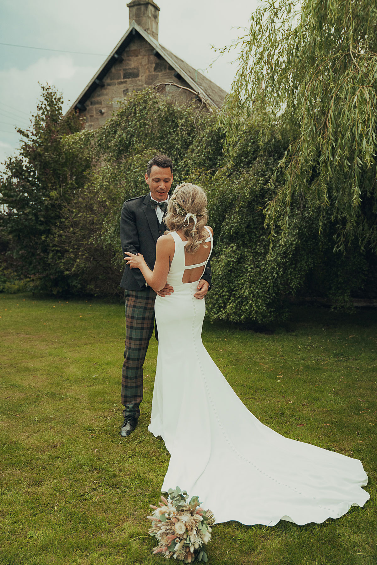 Bride in a fitted sleeveless wedding dress with button detail and low back stands with groom in tartan trews outdoors beside greenery and a stone building, her bouquet placed on the grass in the foreground