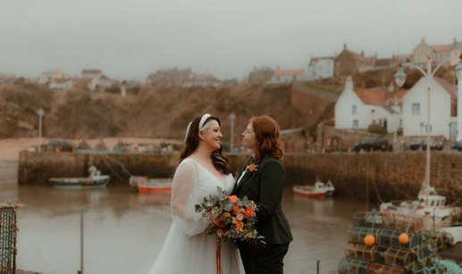 two brides, one wearing a wedding dress, one wearing a suit, look into each others' eyes with Crail and harbour in the background