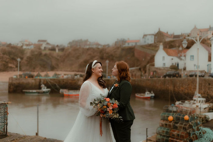 two brides, one wearing a wedding dress, one wearing a suit, look into each others' eyes with Crail and harbour in the background