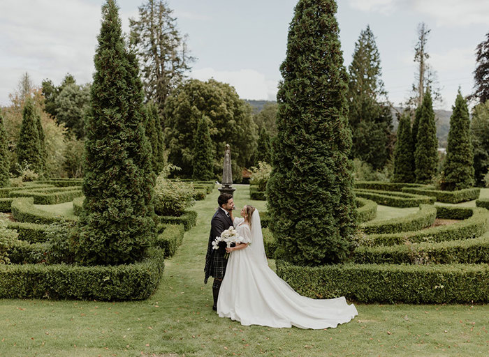 a bride and groom pose for a photo amid sculptured hedges and tall trees in the garden at Achnagairn Castle