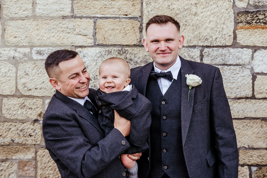 Two men stand holding a baby, all wearing dark blue suits