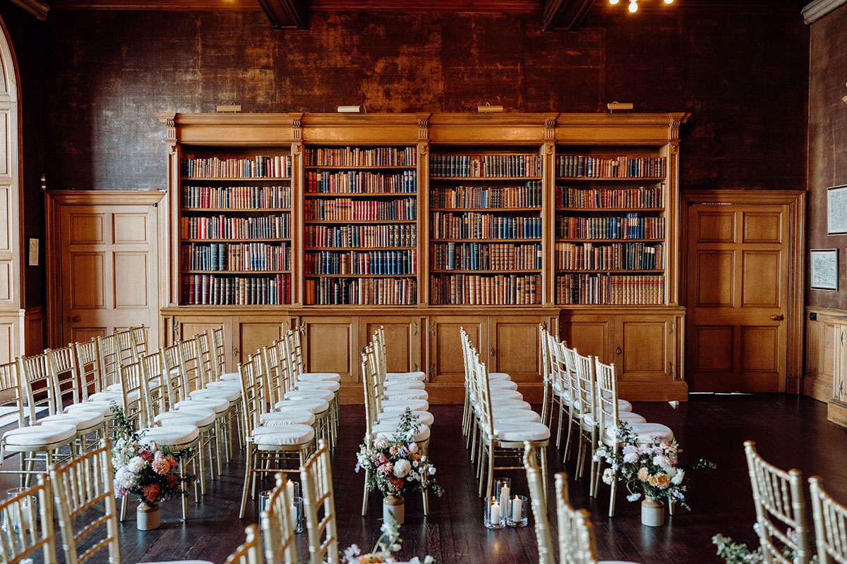 Wedding ceremony setup inside Barnbougle Castle library featuring gold chiavari chairs, floral aisle arrangements and floor-to-ceiling bookshelves