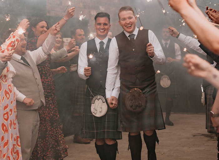 Two grooms holding hands and also holding sparklers wearing Highlandwear smiling whilst walking through a crowd of guests