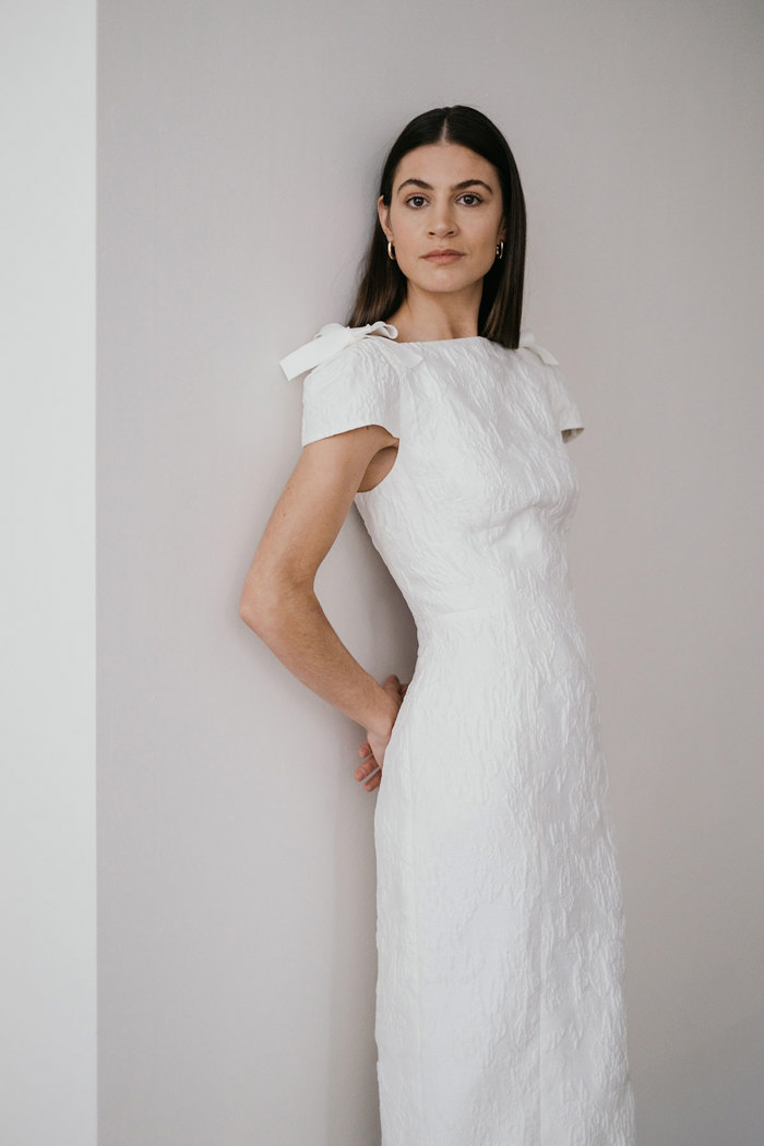dark haired model leans against a plain white wall with her hands tucked behind her back as she wears a white textured column wedding dress with a small bow placed on the top of each shoulder