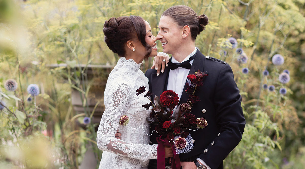 Bride in a high-neck lace wedding dress holding a burgundy bouquet and smiling with the groom in a black tuxedo in the gardens of Carlowrie Castle near Edinburgh.