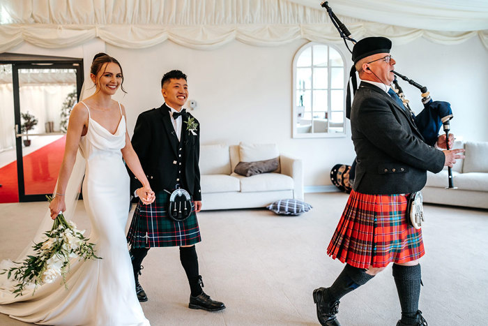 bride and groom hold hands walking into marquee at mar hall as piper wearing red tartan kilt leads them in