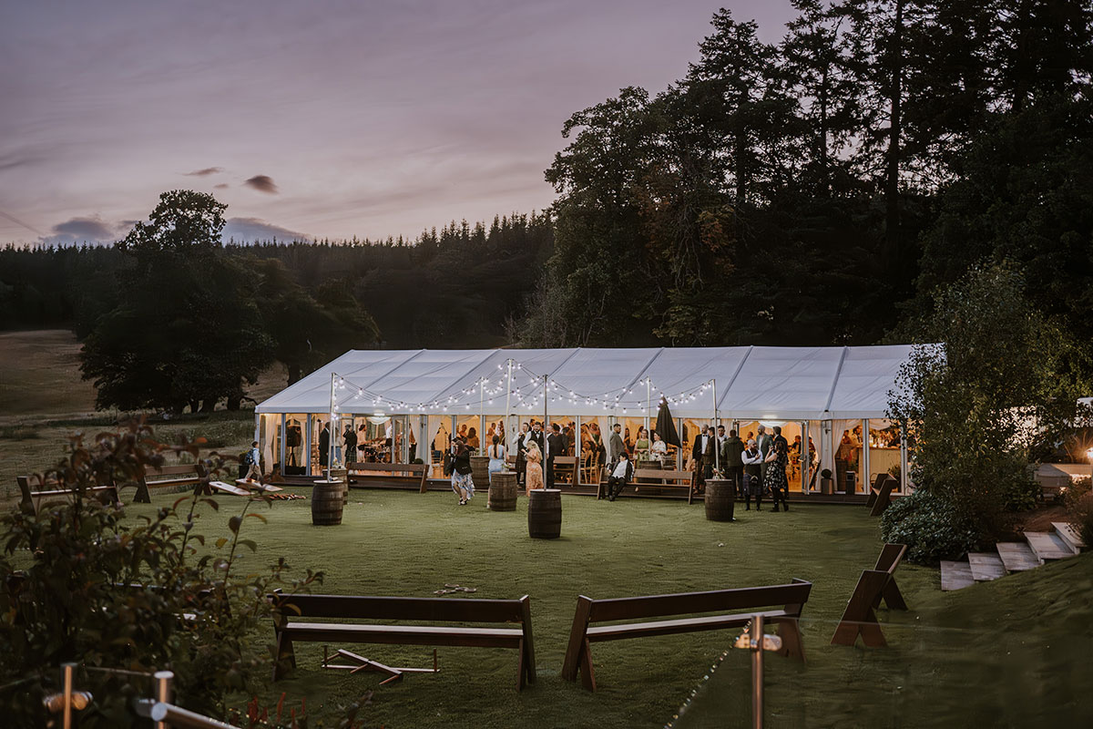 Evening wedding reception in a clear-roofed marquee with string lights and guests mingling on the lawn outside