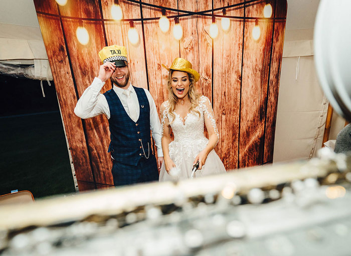 Bride and groom wear prop hats in front of photobooth screen made to look like wooden paneling