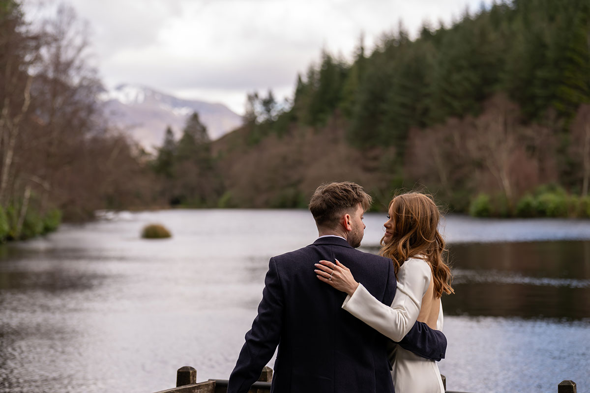 Bride and groom embracing beside a peaceful loch after their Glencoe elopement, with Scottish Highland scenery in the background.