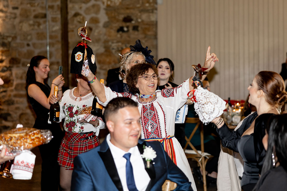 Wedding guests in traditional Romanian clothing lead a lively celebration inside Falside Mill, holding symbolic items and champagne.