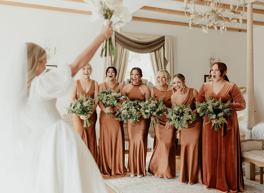 six bridesmaids wearing rust-coloured dresses hold bouquets, laugh and look surprised as a bride in foreground holds bouquet aloft. They are in an elegant room decorated in cream with light wooden details and draped curtains in the background