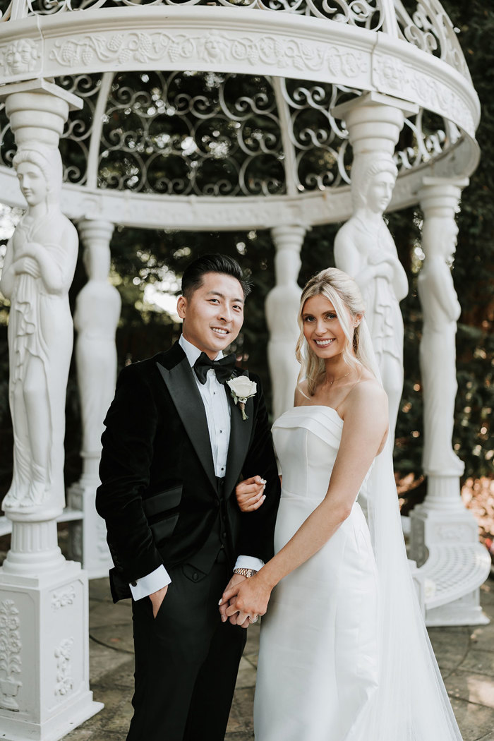 a bride and groom posing in front of white ornamental pergola in garden at Busby Hotel