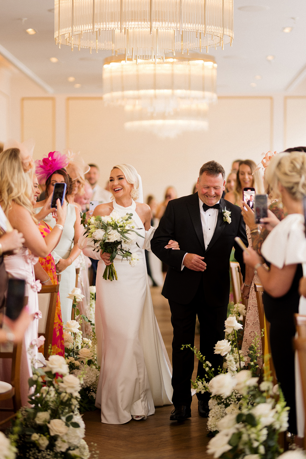 Bride walking down the aisle with her dad as guests take photos during the ceremony