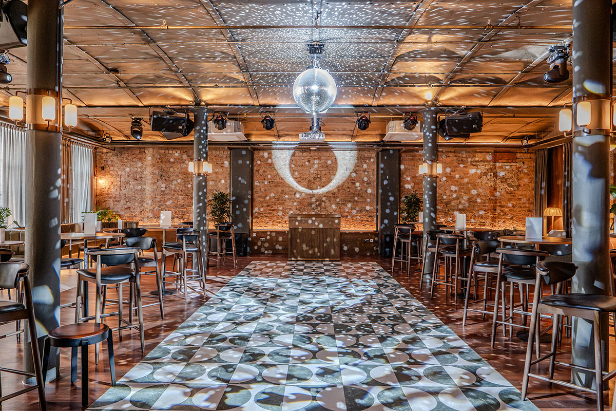 A large event room with a dancefloor, exposed brick walls and a central disco ball casting light patterns.