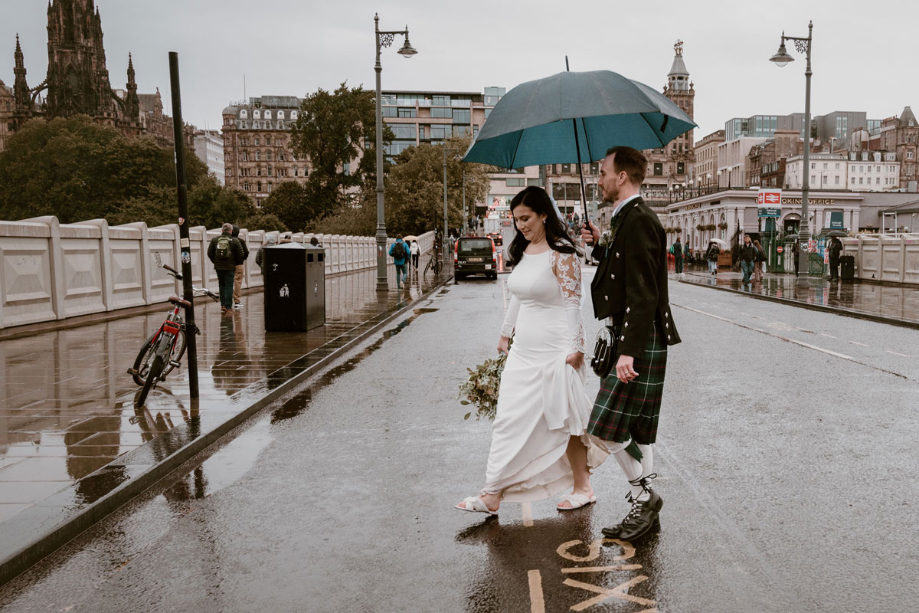 Groom holds umbrella over bride as they walk through Edinburgh