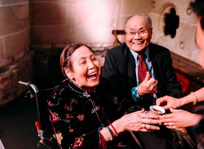 a seated person being given a small cup of tea as another happy person looks on