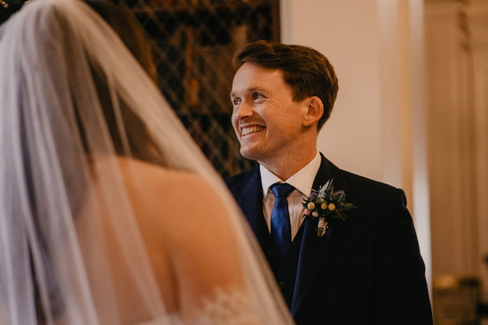 A Person Wearing A Blue Tie And Buttonhole Smiling With A Bride Wearing A Veil In Foregroun